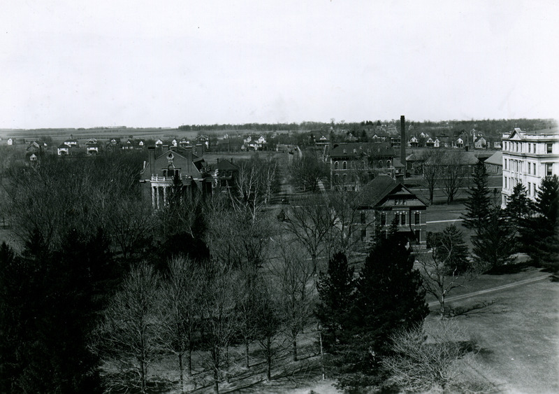 In this view looking west, Alumni Hall is on the left, Chemical and Physical Laboratory is to the left of the smoke stack, and the light-colored building on the right is Engineering Hall II (later known as Marston Hall). A man is attending to his horse on the unpaved street in the center of this view and there are rows of homes in the background, 1913.