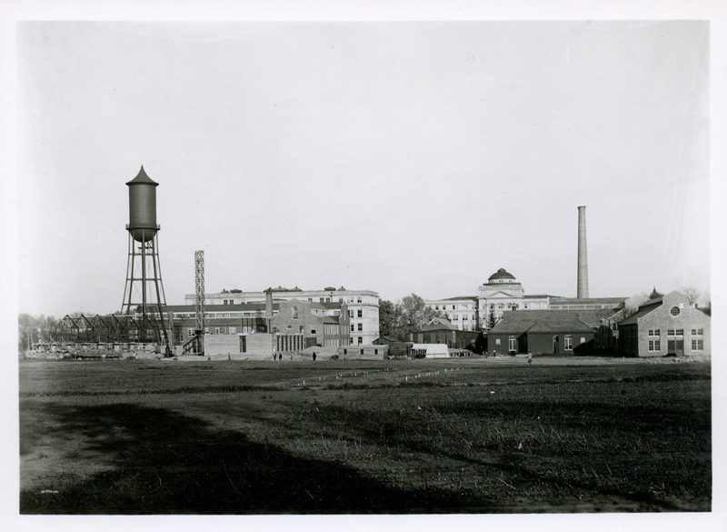 Workmen are constructing the Industrial Arts and Engineering Experiment Station, adjacent to the Marston Water Tower. People are walking by the construction site, 1914.