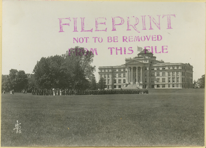 Graduates, attired in their robes, are in a procession walking across campus.There are only a few women in the graduating class. One woman is carrying a parasol. A man on horseback is in front of Central Building (later known as Beardshear Hall), 1906.