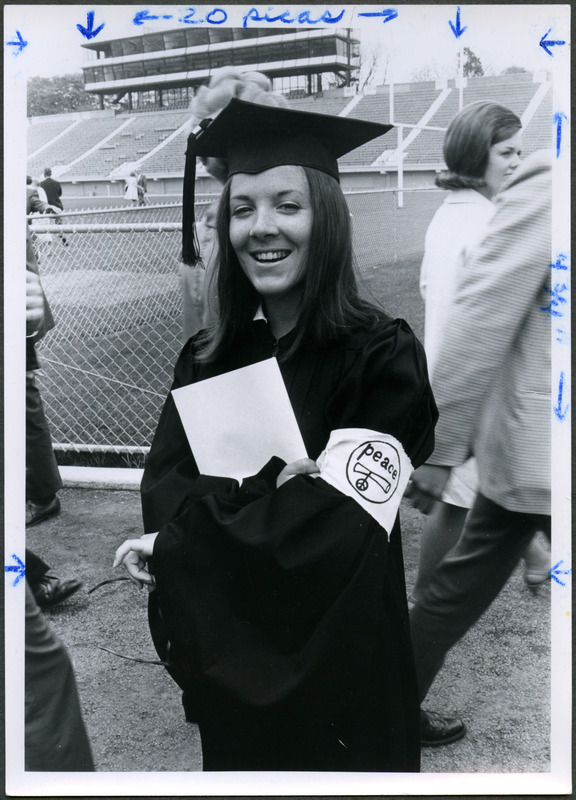 A female graduate is standing in front of Clyde Williams Field displaying a peace decale on the sleeve of her robe, 1970.