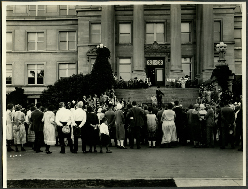 Members of the senior class and alumni are gathered in front of Central Building (later known as Beardshear Hall) listening to a man speaking. Several members of the audience are laughing and appear to be enjoying the talk, 1925.