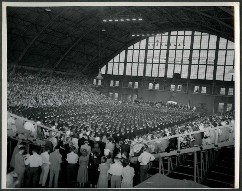 Graduates are sitting on the main floor of the Armory during the commencement ceremony. Family and friends are are seated in the bleachers, 1949.