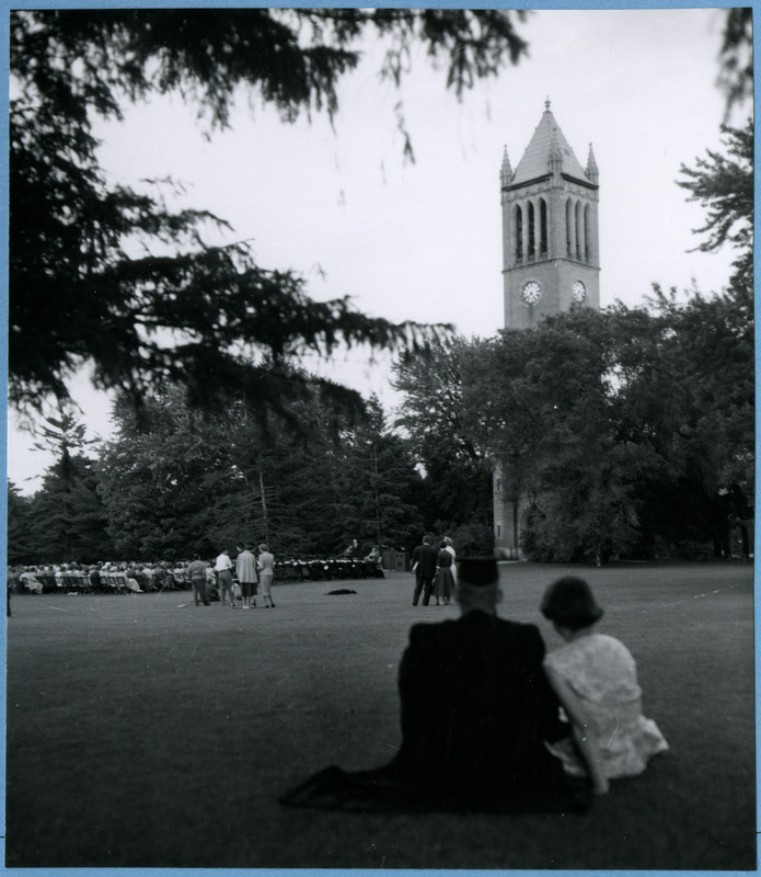 Graduates are seated in chairs near the Campanile for the Cap and Gown Ceremony. Families and friends are seated behind them, 1955.