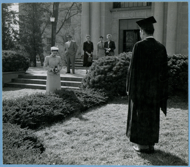 A woman is taking a photograph of a male graduate in front of the Memorial Union. A man and three other women are looking on, 1957.