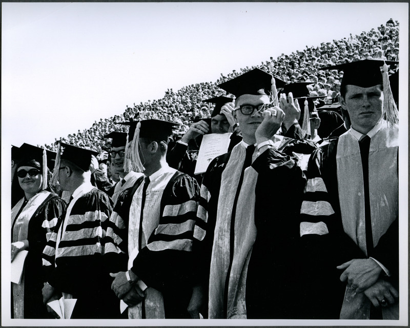 Graduates, attired in cap and gowns, are standing for graduation ceremonies in Clyde Williams Field, 1969.