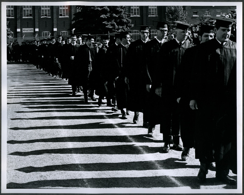 Graduates are in a procession walking from State Gymnasium to Clyde Williams Field for commencement ceremonies. There is a "Reception" banner hanging on State Gymnasium, 1969.