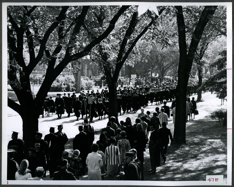 People are on the sidewalk watching the graduates walking in a procession along the street, 1969.