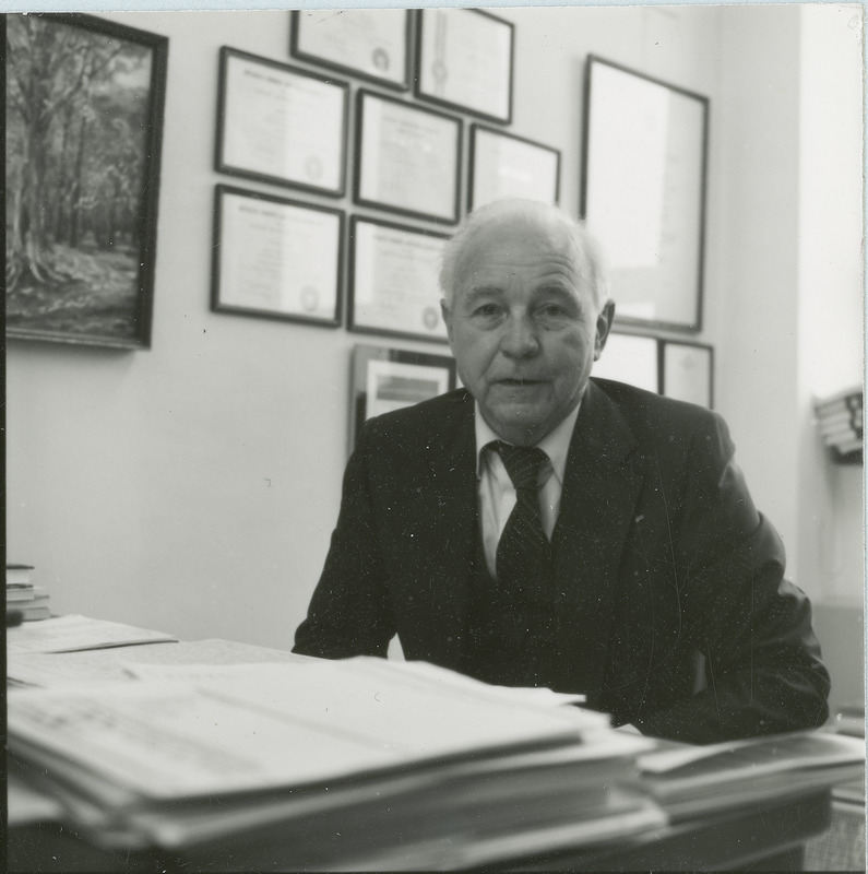 Earl O. Heady, Distinguished Professor of Economics, is sitting at the desk in his office with papers in the foreground and framed documents on the wall in the background, 1982.