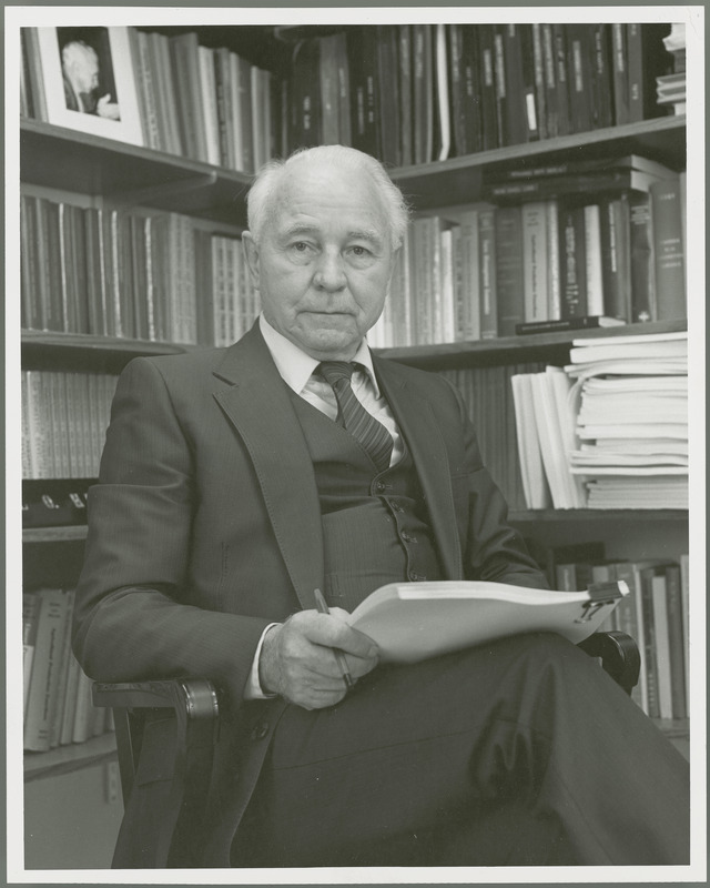 Earl O. Heady, holding papers, is seated in front of shelves of books, 1982.