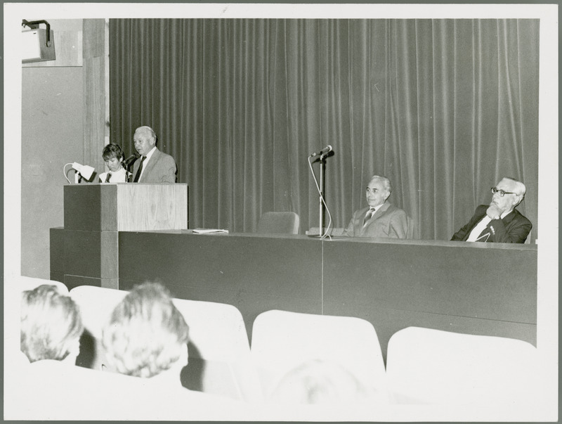 Earl O. Heady, viewed from the right, stands at a lectern speaking before a group in Hungary, with a woman translator on the left and two other men on the podium on the right.