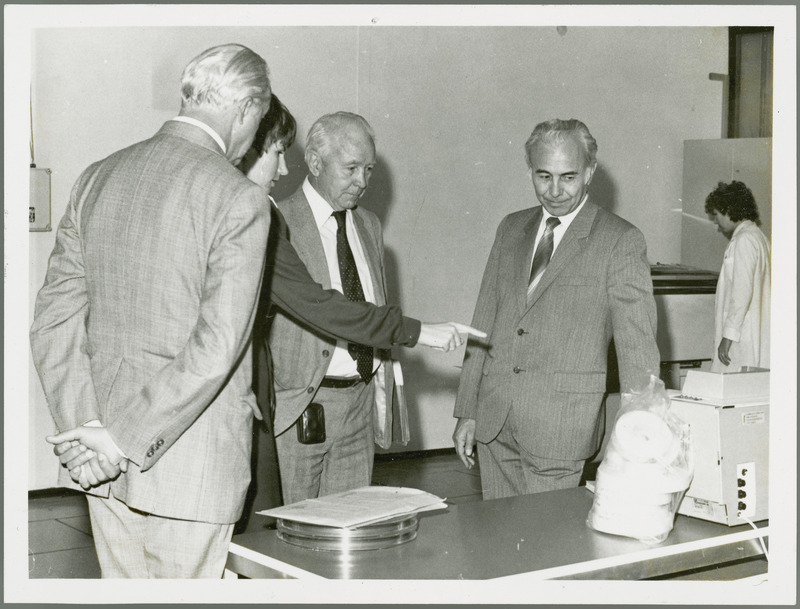 Earl O Heady is being shown research equipment in Hungary by the woman translator, with J. Toth and another man, 1979. Tape reels and rolls of white tape appear to be related to computers, and a woman in the background watches a machine.