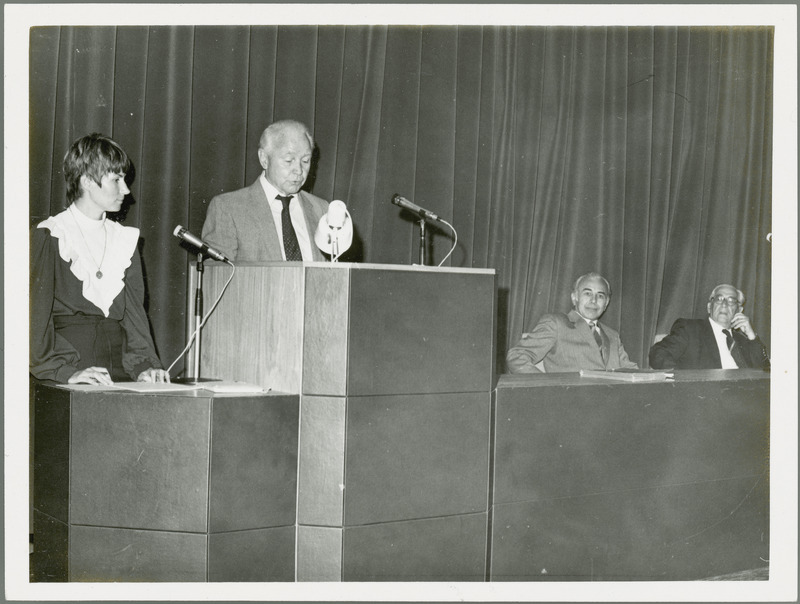 Earl O. Heady, viewed from the left, stands at a lectern speaking before a group in Hungary in 1979, with a woman translator on the left and two other men on the podium on the right.