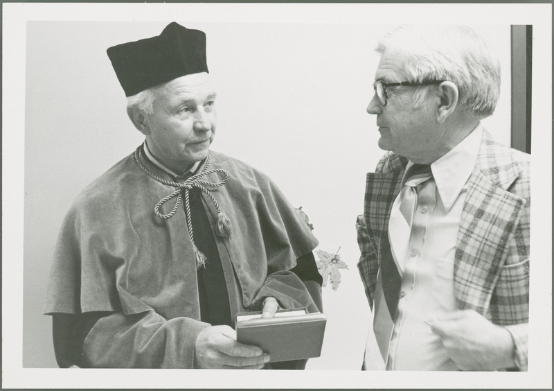 Earl O. Heady, with title Benela, wears the Polish academic robes he received as part of an honorary degree from the Warsaw Agricultural University in 1979. He is holding an award case, while talking with Department of Economics Chair Raymond Beneke.