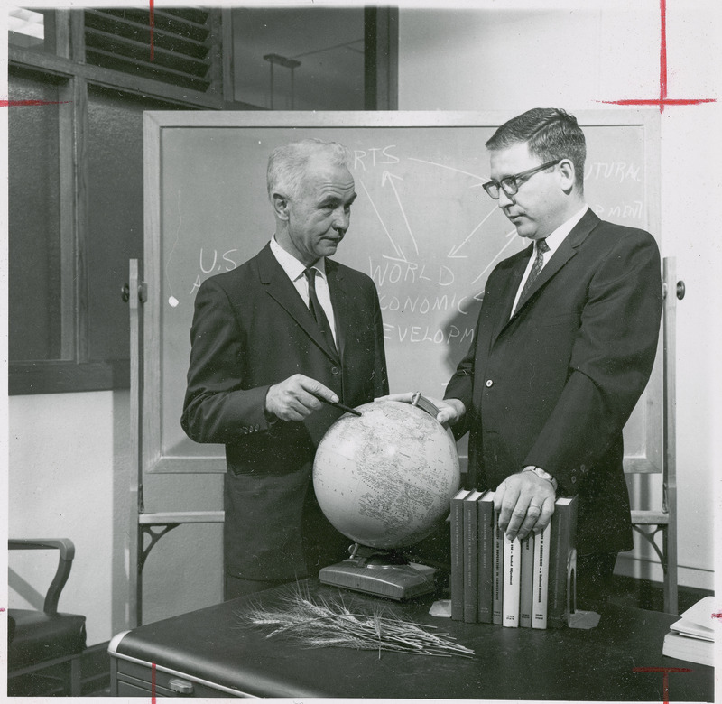 Professor Earl O. Heady points at a globe while talking with Associate Professor Leo V. Mayer, whose right hand is on the globe and his left hand rests on a set of Iowa State agricultural economics books. Some wheat stalks lie on the desk in the foreground, while the blackboard behind the men has a diagram about world economic development.