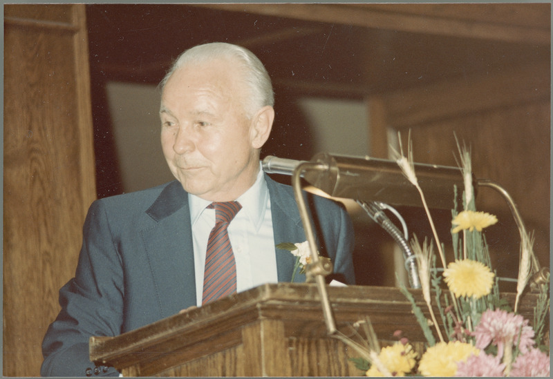 Earl O. Heady, wearing a boutonniere, looks to his right with amusement while standing behind a lectern with yellow and pink flowers in front of it.