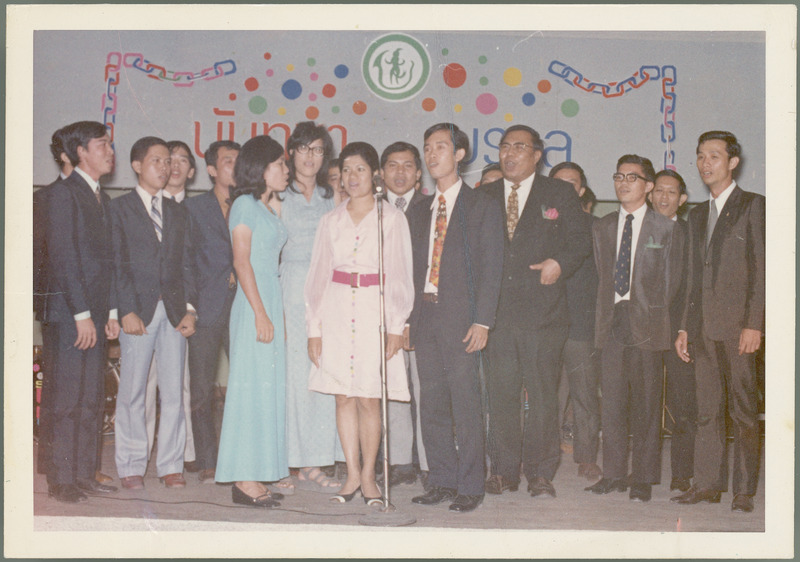 A group of Indonesian students in suits and dresses sing in front of a microphone with a festive backdrop of colorful links, bubbles and Indonesian and English lettering. This was apparently for Earl O. Heady, Nov. 20, 1972.