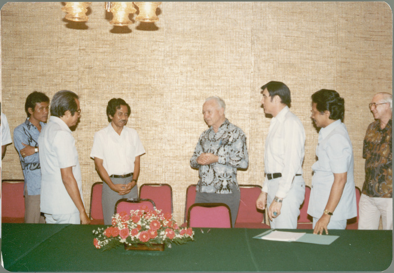 Earl O. Heady stands in the center of the group of Indonesian and American collaborators behind a green-covered table on which the Indonesian Project Contract rests. The table has a centerpiece of red and white flowers, and red banquet chairs are lined up along the wall in the background.