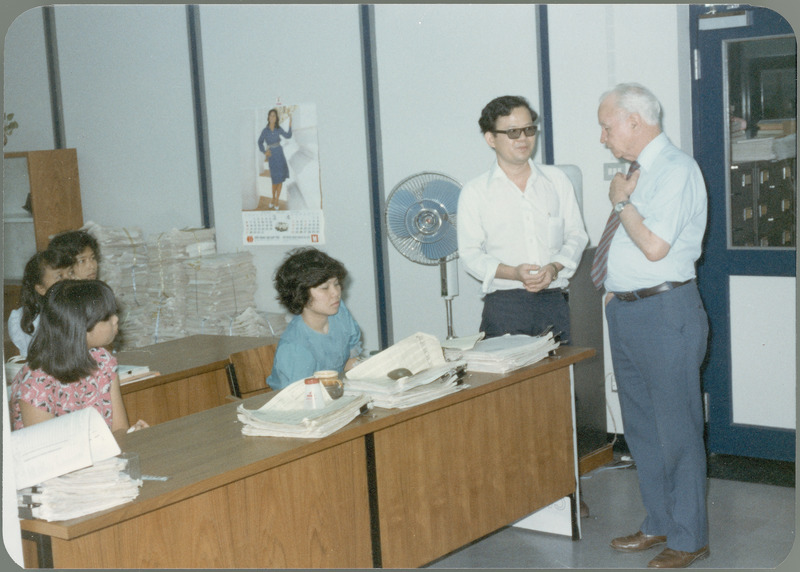 Earl O. Heady talks with an officer at the Ministry of Agriculture, Office of Agricultural Economics, in Bangkok, Thailand, April 1983. On the left, four young women sit at desks with computer printouts, while more printouts are stacked in the background under a calendar by a large fan.