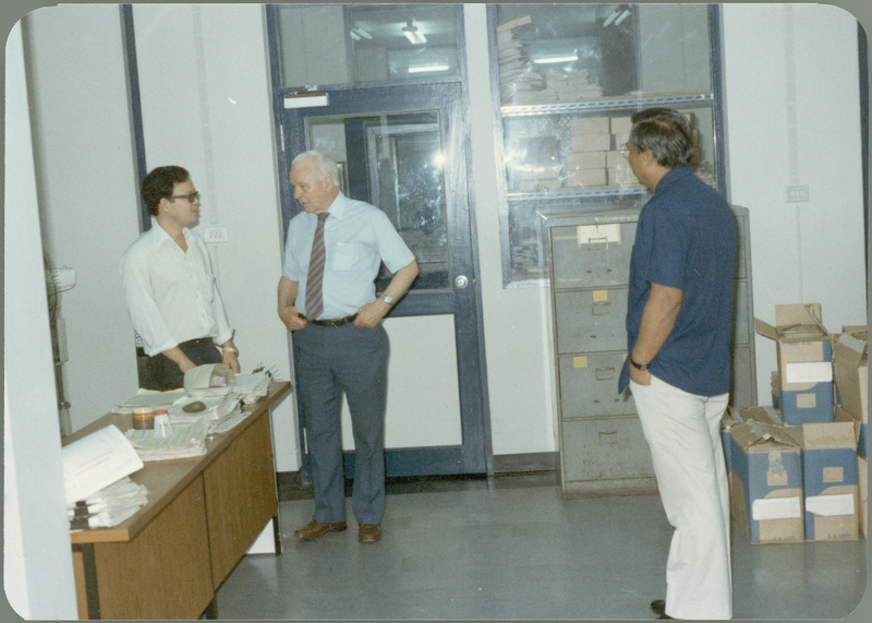 Earl O. Heady talks with blue-shirted Somnuk Sriplung, chief, Office of Agricultural Economics in the Ministry of Agriculture, and another official, in one of their office areas, Bangkok, Thailand, April 1983. Grey file cabinets, boxes of supplies, and the door and window to another office area are visible in the background.