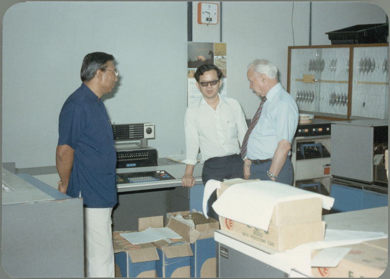 Earl O. Heady hears about upgraded equipment at the Office of Agricultural Economics computer center with office chief Somnuk Sriplung and another official who is explaining it, April 1983. Boxes of supplies are on the floor and a table, and storage for computer reels is on the right.