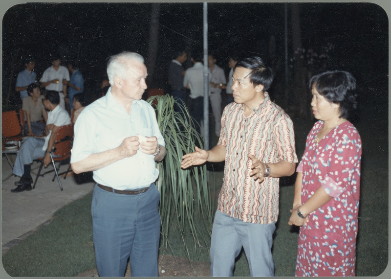 Earl O. Heady stands outside at an evening social event in Thailand talking with a man and woman, with other people talking together in the background, April 1983.