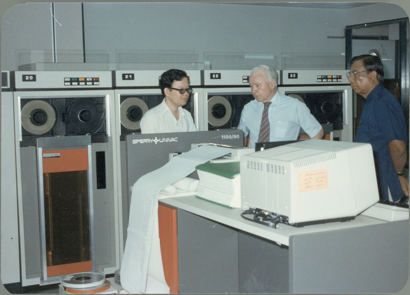 Earl O. Heady hears about upgraded equipment at the Office of Agricultural Economics computer center in Bangkok, Thailand, with office chief Somnuk Sriplung and another official who is explaining it, April 1983. The new Sperry/Univac 1100/60 mainframe is part of the equipment shown, with a printer operating in the foreground.