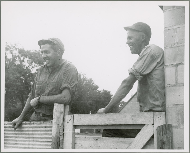 Two smiling young men wearing caps, with their shirt sleeves rolled up, lean on a fence made of corrugated metal with a wooden gate.