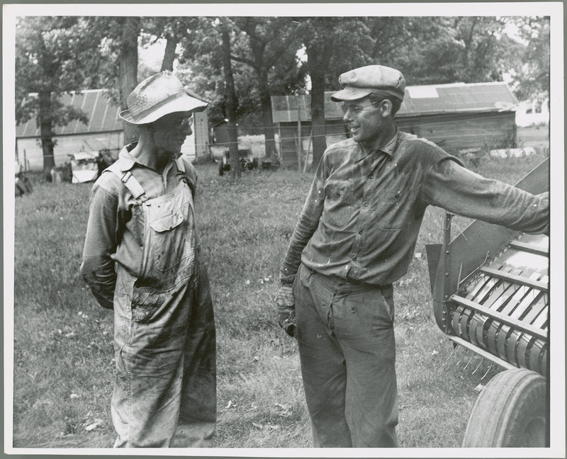 Two men who look dirty from their hard work stand talking by a farm machine, perhaps a hay baler, with two one-story farm buildings in the background with trees. Both men wear hats and glasses, and the older man wears overalls.