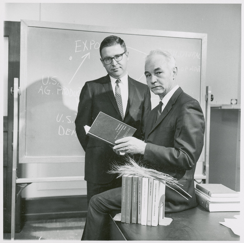 Associate Professor Leo V. Mayer stands by Earl O. Heady, director of the Center for Agricultural and Economic Development, as CARD was called at that time. Dr. Heady sits on a desk and holds open a book entitled Alternatives for Balancing World Food Production Needs. Other books about agricultural economics are lined up on the desk, covered by a few stalks of wheat, and a blackboard showing a diagram about agricultural production is in the background, 1966.