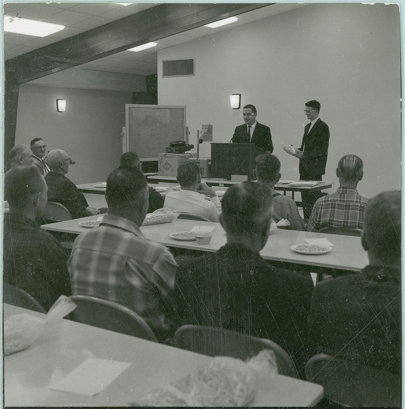 A man in a suit presents an agricultural education lecture for a class of adult men, 1966. The class sits on folding chairs at folding tables, with bags of corn kernels or soybeans in front of them as examples. The lecturer stands behind a lectern on a table, with a portable blackboard, a slide projector, and an overhead projector to his right. A young man in a suit stands by him holding a bag of corn or soybeans.