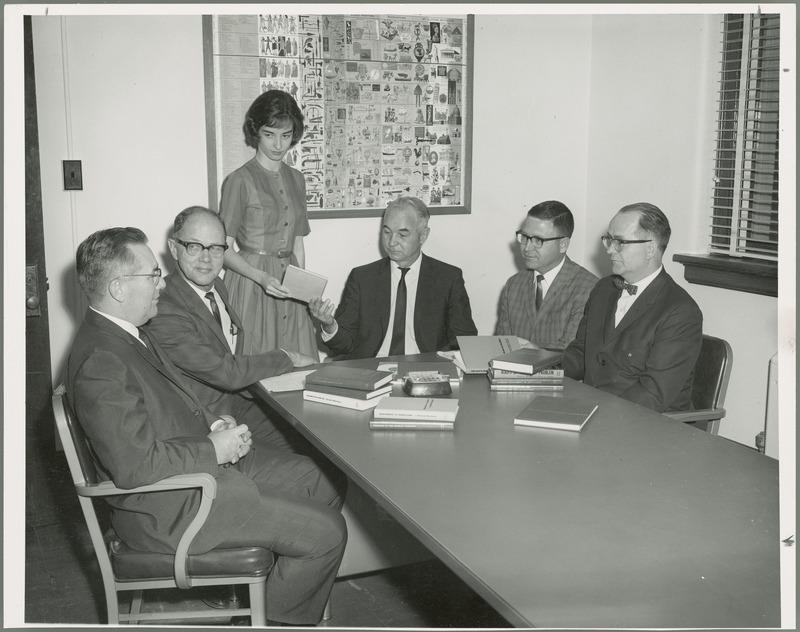 Key figures in the Center for Agricultural and Economic Development (as CARD was then called) are seated around a table with books, and a poster of the achievements of civilization through the centuries on the wall in the background, 1966. They are, from left: William G. Stucky, education leader; Wallace E. Ogg, subject matter coordinator; Mary Romero, secretary; Earl O. Heady, executive director; Leo Mayer, associate, and Edwin O. Hardolsen, editor. Mary Romero is handing Earl Heady a book, but he is looking down and everyone else is looking at William Stucky.