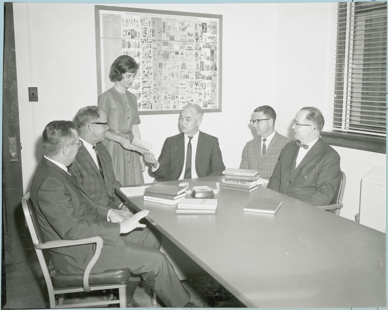 Key figures in the Center for Agricultural and Economic Development (as CARD was then called) are seated around a table with books, and a poster of the achievements of civilization through the centuries on the wall in the background, 1966. They are, from left: William G. Stucky, education leader; Wallace E. Ogg, subject matter coordinator; Mary Romero, secretary; Earl O. Heady, executive director; Leo Mayer, associate, and Edwin O. Hardolsen, editor. Mary Romero is handing Earl Heady a book, and everyone is looking at Earl as he speaks.