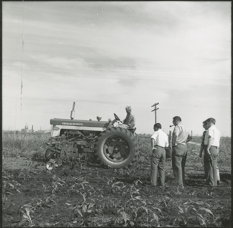 Four men observe a tractor with extra equipment performing work on the soil in a field, for a Rural Minister Short Course, 1961.