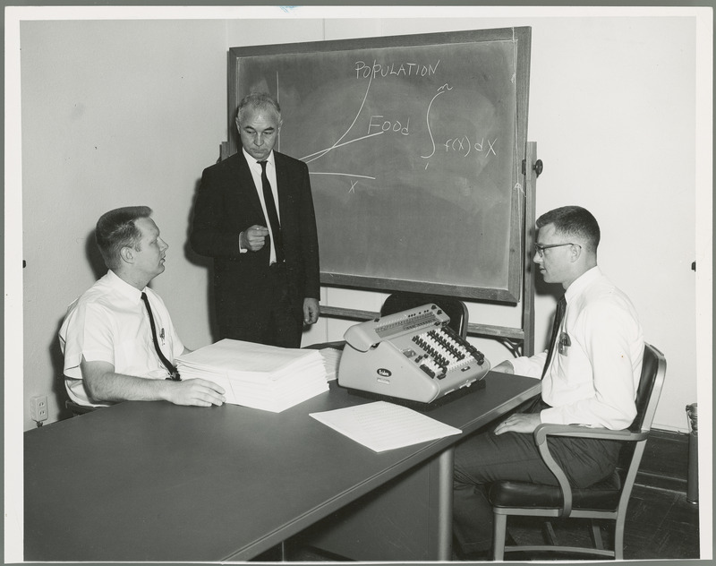 Earl O. Heady stands at blackboard, while Gaylord Worden sits on the left with a stack of printouts, and Leroy Blakeslee sits on the right with a calculating machine. All of them are part of the Center for Agricultural and Economic Development, 1966.