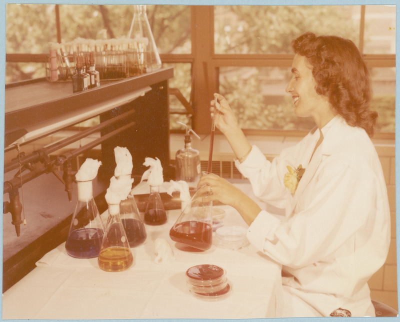 A woman researcher transfers colored fluids between beakers and petri dishes with pipette.