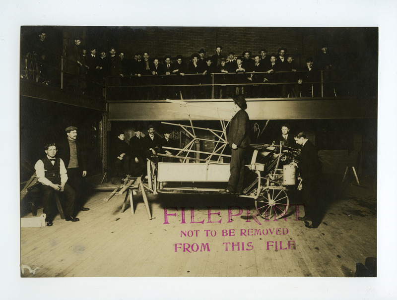 An instructor lectures a group of men on a pulley driven reaper. Some men are standing around the reaper, while other observe from a balcony.