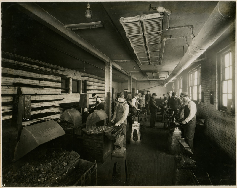 A group of students are working in the Forge Shop. They are using forges, anvils and hammers to shape the hot metal. One of the forges is labeled, "Buffalo Forge Co., Buffalo, NY.".