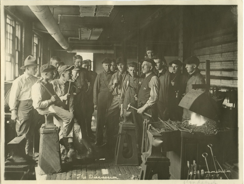 An instructor in the Blacksmithing class is demonstrating a technique. He stands behind an anvil with a hammer in one hand and a piece of metal in the other.