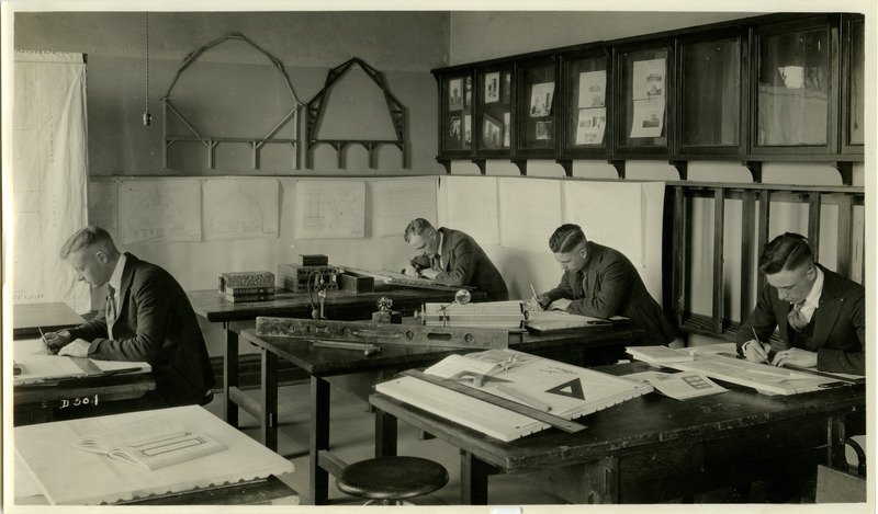 A group of students are seated at tables working at drafting projects. Various pieces of drafting equipment are on the tables including right angle triangles, levels and a Thacher's Calculator (cylindrical slide rule).