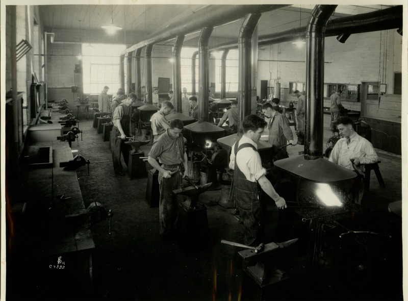 A group of men are working at individual work stations in the Forge Shop. Some men are heating the metal in the forge, while others are shaping it on the anvil with hammers.