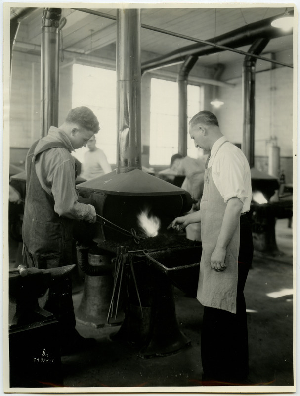 A man is using a set of tongs to heat a piece of metal in the forge. An instructor offers advice on the process.