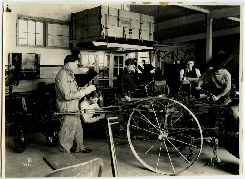 A group of men are examining a cultivator, perhaps manufactured by the Harrow Company. The cultivator is attached to a wheeled mechanism which is labeled: Manufactured by the Light draft Harrow Company.