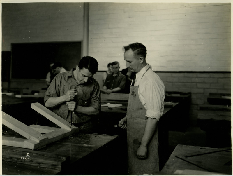 A man is tightening a screw on a saw horse type object that he is constructing upon a work bench. An instructor looks on while other students work on projects in the background.