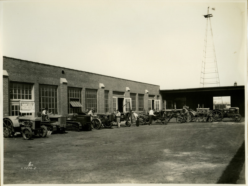 In an outdoor courtyard, a group of men are examining various types of tractors.