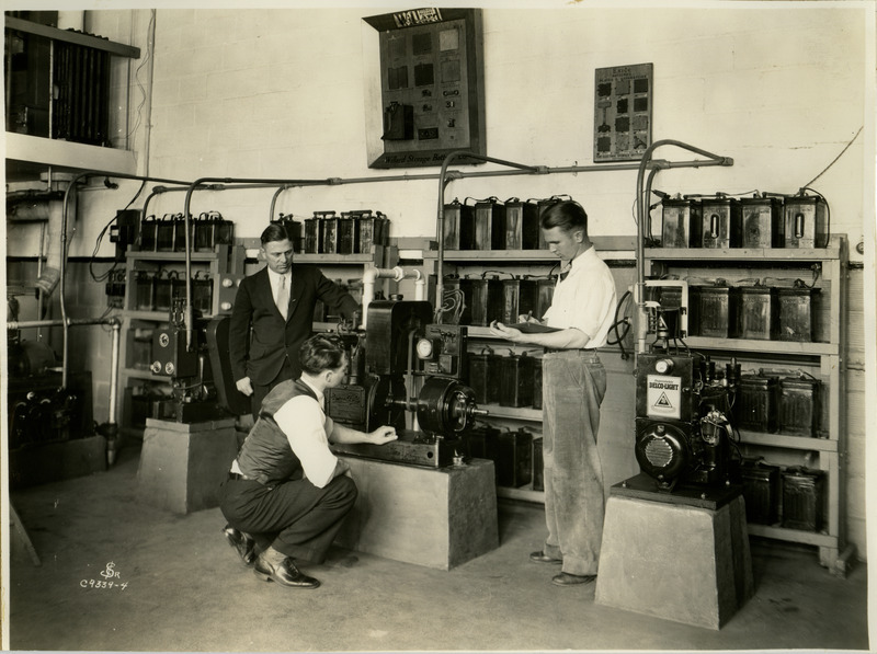 A group of three men are examining batteries and other equipment designed to bring electricity to farms.