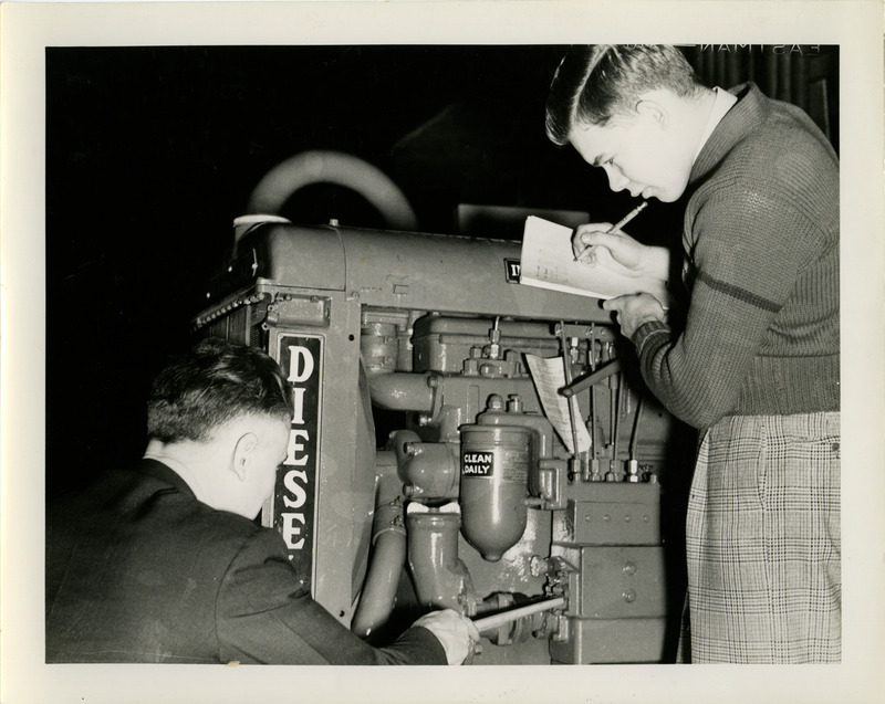 Two men examine the engine of a diesel tractor. One man is pointing out parts, while the other is taking notes.