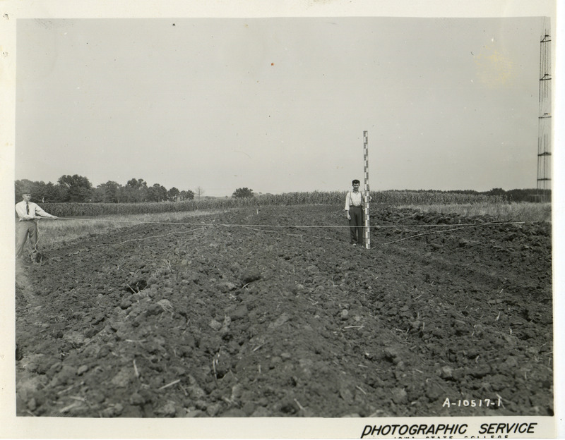 Two men are standing in a plowed field. One is holding a measuring rod while the other users a stretched rope to indicated the depth of the furrow.