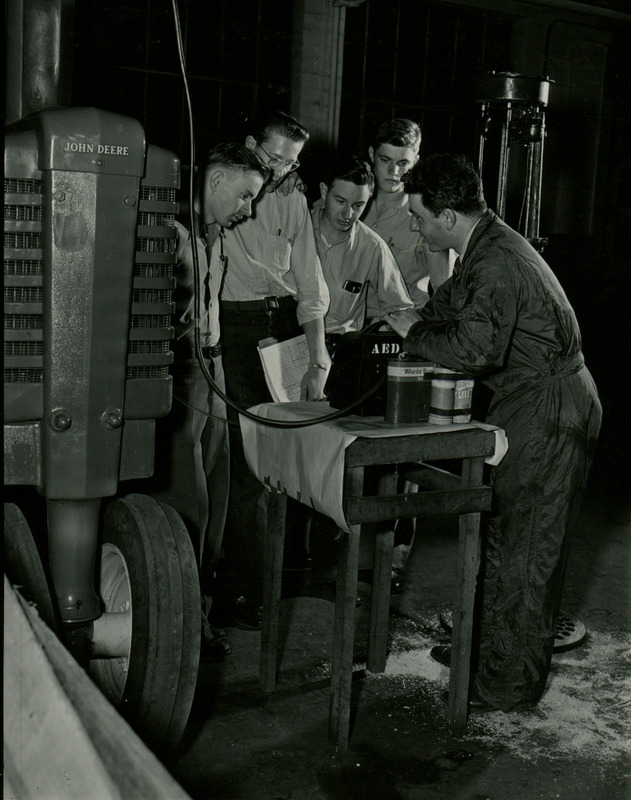 Four men listen to an instructor explaining a tractor carburetor. The group of men are standing next to a John Deere tractor.