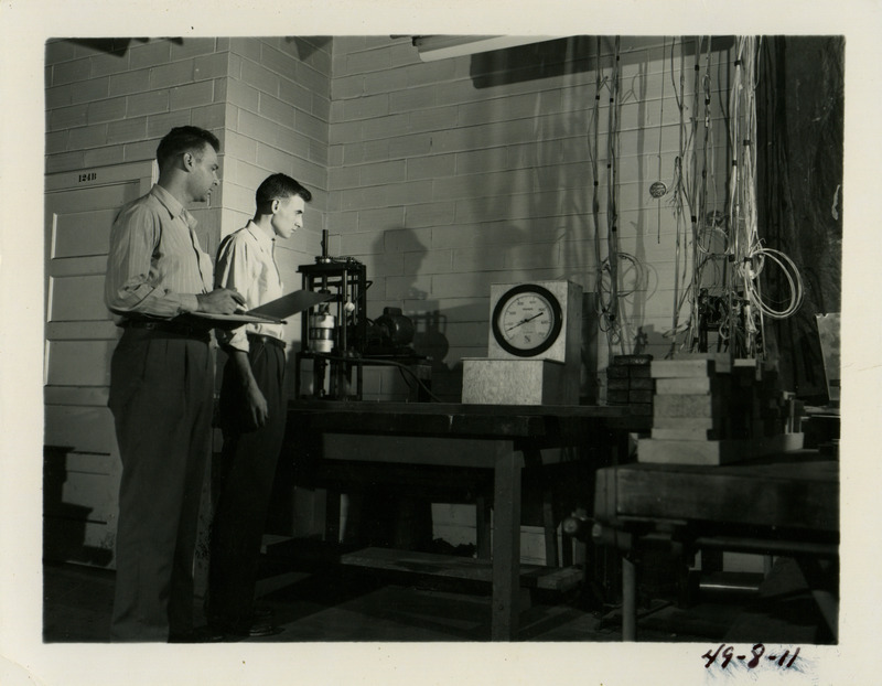 Two individuals observe nail testing equipment. One individual is holding a clipboard on which he is probably recording the results of the test.