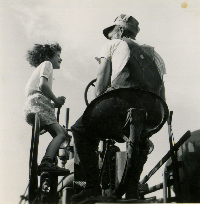 Little Sorenson rides on a tractor by standing on the side with Irving Sorenson driving, showing a farm safety issue.